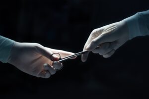 A hand with a plastic glove handing a surgical scissor over to someone else wearing a plastic glove.